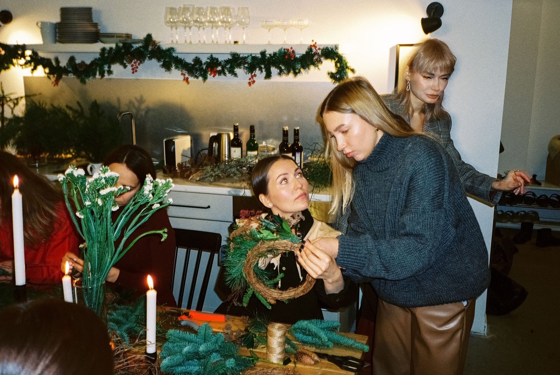 Three people decorate a Christmas wreath at a candlelit bar, with garlands and bottles in background.